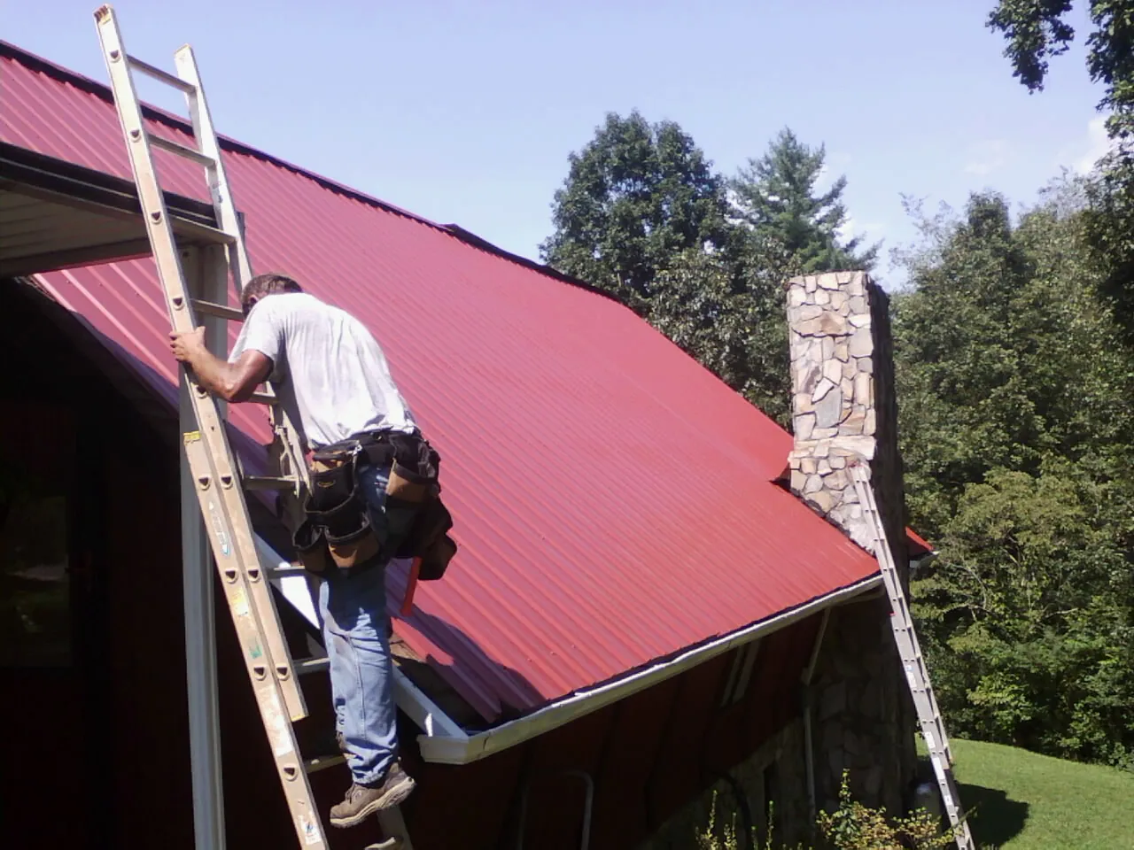Roofer on a ladder beside a red metal roof and stone chimney — Soffit & Fascia Repair by Roofing Brothers in Roanoke